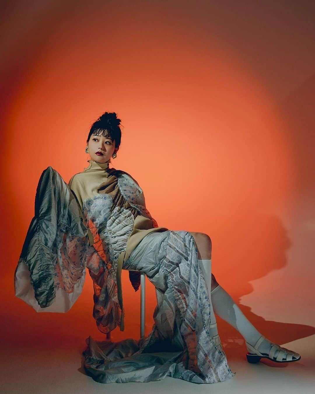 Model seated against a red-orange backdrop wearing a layered, deconstructed kimono